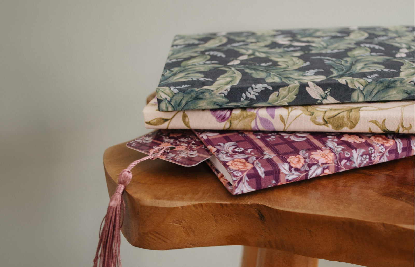 Three stacked fabric book covers or wallets with floral patterns in green, beige, and purple, resting on a rustic wooden stool against a neutral background.
